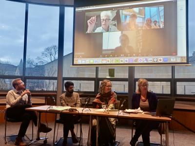 Image courtesy Dr Erika De Vivo Four academics sitting at a desk with microphones, and more scholars on a Zoom screen behind them