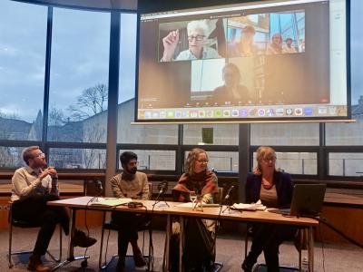 Four academics sitting at a desk with microphones, and more scholars on a Zoom screen behind them